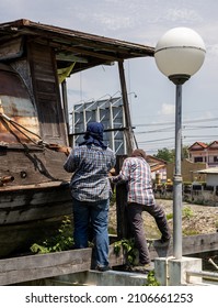 A Group Of Thai Male Workers Are Installing Large Rope Slings To Lift And Move An Old Wooden Boat Moored On Top Of A Canal In A Thai Municipal Market.