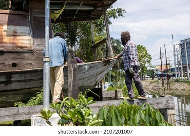 A Group Of Thai Male Workers Are Installing Large Rope Slings To Lift And Move An Old Wooden Boat Moored On Top Of A Canal In A Thai Municipal Market.