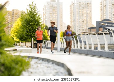 Group of three active multinational people in sport clothes running together outdoors on bridge