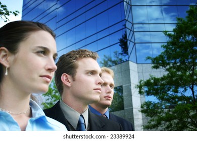 A Group Of Three Business People Standing Together Outside Against A Blue Business Building In Their Business Suits And Business Clothes