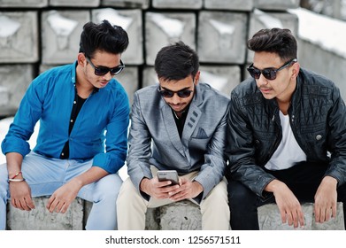 Group of three casual young indian mans in sunglasses posed against stone blocks and looking at mobile phone.
