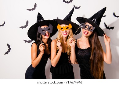 Group of three diverse charming coquettes in dark masquerade elegant dresses, masks on eyes, smiling, enjoying near decorated wall with bats, toothy beaming smiles, scary bright cosmetics hot figures