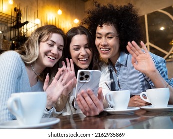 Group Of Three Diverse Young Female Friends Waving Hello On A Video Call With A Smartphone In A Pub