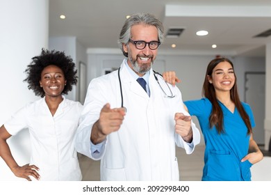 A  Group Of Three Doctors And Nurses Standing In A Hospital Corridor, Wearing Scrubs And Coats. The Team Of Healthcare Workers Are Staring At The Camera And Smiling