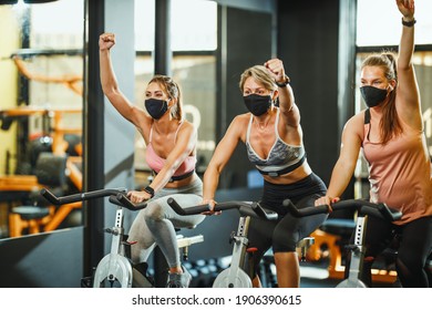 Group Of Three Fit Female Friends With Protective Mask Having Sports Training On Exercise Bikes In A Gym During Covid-19 Pandemic.