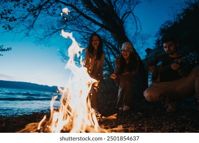 A group of three friends gather by a campfire, laughing and enjoying music played on a guitar by the lake at twilight. - Powered by Shutterstock