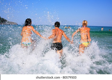 Group Of Three Friends - Man And Women - On The Beach Having Lots Of Fun In Their Vacation In Greece