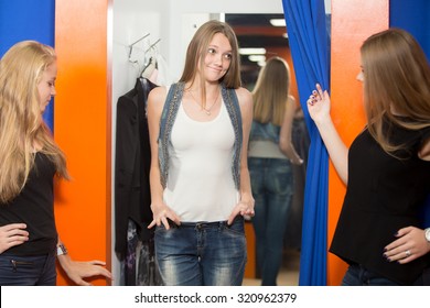 Group Of Three Girlfriends Shopping Together. Cute Young Beautiful Woman Wearing Jeans And Denim Vest Standing In Fitting Room, Pulling A Face, Showing Friends That Her New Outfit Doesn't Fit