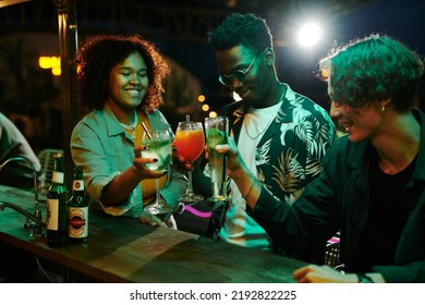 Group of three happy young intercultural people clinking with glasses of alcoholic cocktails by counter while enjoying party in night club - Powered by Shutterstock