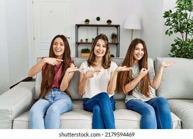 Group Of Three Hispanic Girls Sitting On The Sofa At Home Amazed And Smiling To The Camera While Presenting With Hand And Pointing With Finger. 