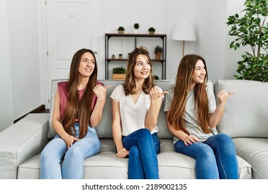 Group Of Three Hispanic Girls Sitting On The Sofa At Home Smiling With Happy Face Looking And Pointing To The Side With Thumb Up. 