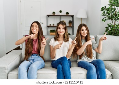 Group Of Three Hispanic Girls Sitting On The Sofa At Home Smiling And Looking At The Camera Pointing With Two Hands And Fingers To The Side. 