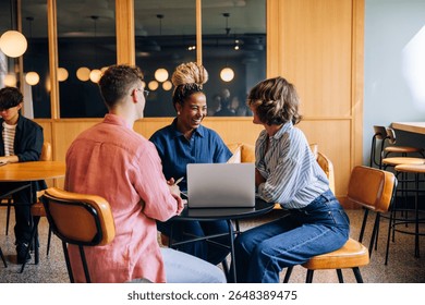 A group of three individuals engage in a constructive conversation around a laptop in a vibrant shared workspace. The atmosphere is professional yet casual, fostering creativity and camaraderie.