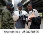 Group of three individuals reviewing captured footage under an urban bridge. Man holding camera, another wearing green jacket and sunglasses, and woman with braids and white mesh top