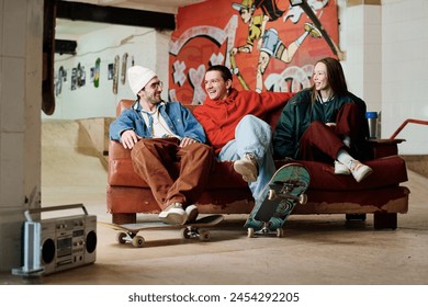 Group of three modern male and female friends hanging out in skatepark sitting on couch, chatting and listening to music, copy space - Powered by Shutterstock