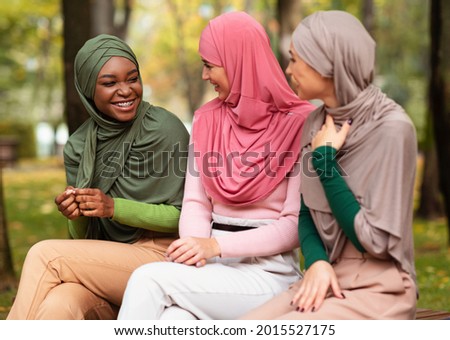 Group Of Three Multiethnic Islamic Women Talking Sitting On Bench In Park Outdoors On Autumn Day, Wearing Hijab. Happy Muslim Females Friends Chatting Enjoying Conversation. Friendship Concept