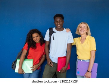 Group Of Three Multi-ethnic Teenage Students With Backpack And Folders On A Blue Wall. Back To School, High School, Diverse Education