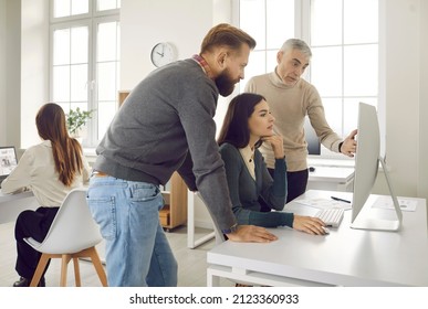 Group Of Three People Looking At Computer Screen And Discussing Something. Team Of Young And Older Corporate Employees Working On Online Project In Modern Light Company Office Interior