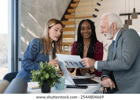 A group of three professionals, including two women and a man of different ethnicities, review documents and engage in conversation at a sleek workplace, indicating teamwork and diversity in business.