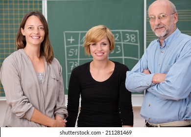 Group Of Three School Teachers With Confident Friendly Smiles Standing In Front Of A Class Blackboard, One Man And Two Women