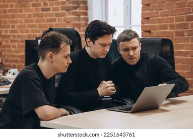 group of three software developers young men in black clothes working with notebook in vintage office with red brick walls, discussion about work