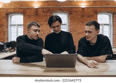 group of three software developers young men in black clothes working with notebook in vintage office with red brick walls, discussion about work