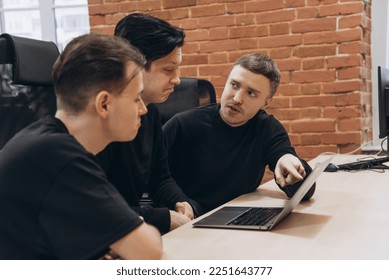 group of three software developers young men in black clothes working with notebook in vintage office with red brick walls, discussion about work