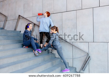 Group of three sports women on the stairs before run. Red head woman drinking water from the bottle, two other stretching and listening music with headphones.