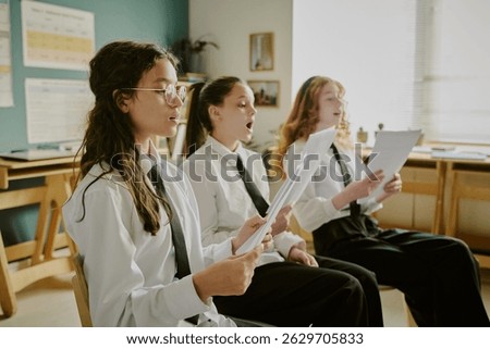 Group of three students practicing singing from sheet music in classroom seating arrangement, focusing on vocal exercises and team coordination