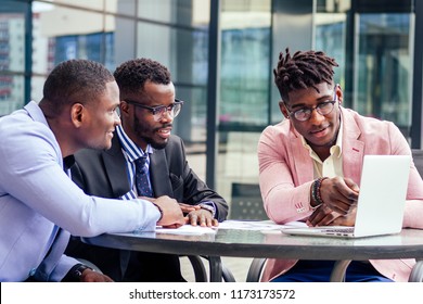 A group of three stylish African American students entrepreneurs in fashion business suits working sitting at table with laptop in a summer cafe outdoors