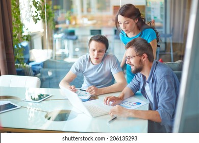 Group Of Three Successful Business Partners In Casual Using Laptop At Meeting In Office