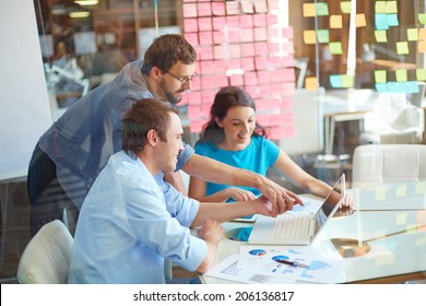 Group Of Three Successful Business Partners In Casual Looking At Laptop Screen While Two Young Men Pointing At It At Meeting In Office