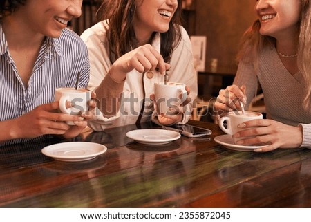 Group of three unrecognizable girl friends drinking coffee sitting in a table of a cafe. They show their friendship while they have fun. We only see their smiles not the rest of the faces.