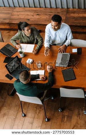A group of three workers sit at a table and hold pens and pencils while one of them talks