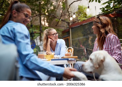 Group Of Three Young Adult Beautiful Caucasian Female Friends Sitting In Outdoor Cafe With A Pet Dog, Talking, Smiling, Laughing