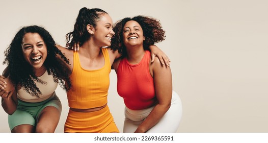 Group of three young, diverse female athletes celebrate their healthy and active lifestyle in a sports studio, smiling and laughing together while wearing sporty fitness clothes.