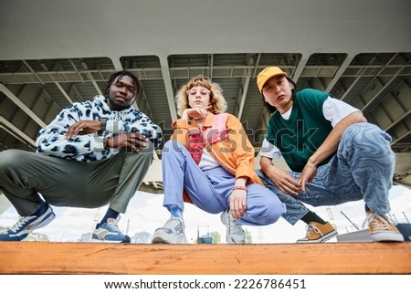 Group of three young people wearing street style clothes outdoors while sitting on stairs in urban area and looking at camera