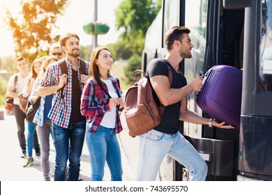 A group of tourists enters the bus. The guy helps the girl to bring the luggage to the bus. They will travel on a modern black bus. Behind them is a group of tourists with luggage.
