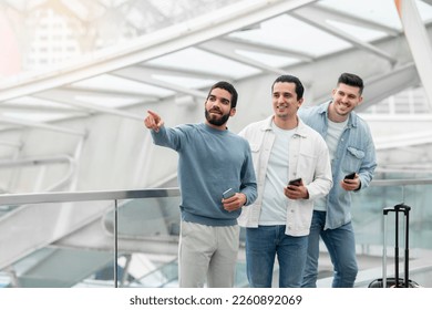 Group Of Tourists Guys Using Phones Pointing Finger Aside Advertising Travel Application Standing With Suitcase In Airport Terminal Indoors. Mobile Services For Tourism Concept - Shutterstock ID 2260892069