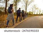 Group of tourists hiking or trekking on warm September day. View from behind team of young people with backpacks walking together on roadside along highway lined with green trees