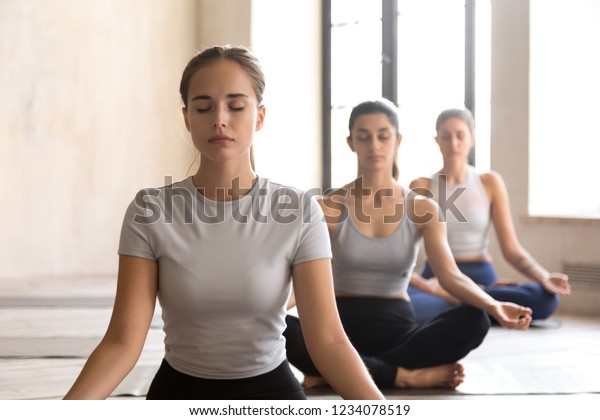 Group training of deep meditation at yoga studio class. Diverse young females sitting with closed eyes in lotus position meditate and visualizing together, feeling self-awareness serenity and calmness