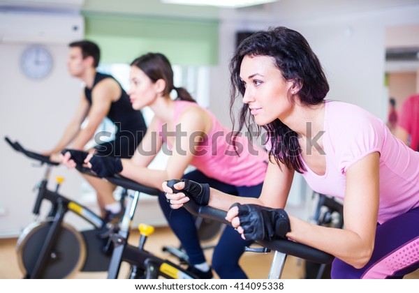Group training people biking in\
the gym, exercising legs doing cardio workout cycling\
bikes.