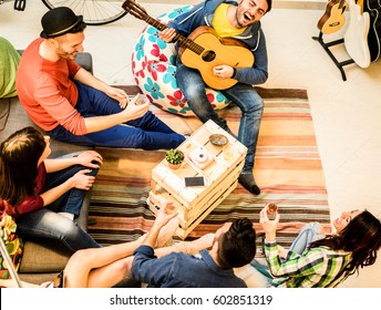 Group of trendy friends having fun in hostel living room - Happy young backpackers enjoying time together playing music with guitar and drinking beer - Focus on musician and wood table - Warm filter