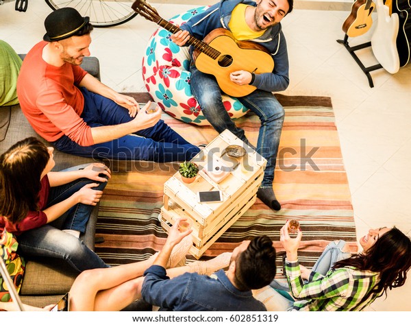 Group of trendy friends having fun in hostel\
living room - Happy young backpackers enjoying time together\
playing music with guitar and drinking beer - Focus on musician and\
wood table - Warm filter