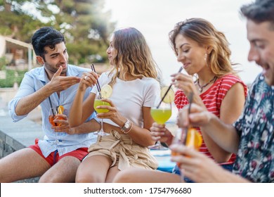 Group of trendy happy young people talking and having fun drinking tropical cocktails together sitting on a bench outdoor in the summer