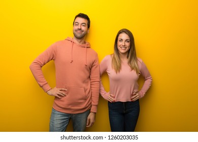 Group Of Two People On Yellow Background Posing With Arms At Hip And Laughing Looking To The Front