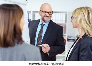 Group Of Two Women And One Smiling Bearded Man With Eyeglasses Meeting And Discussing Something Next To Chart In Business Office