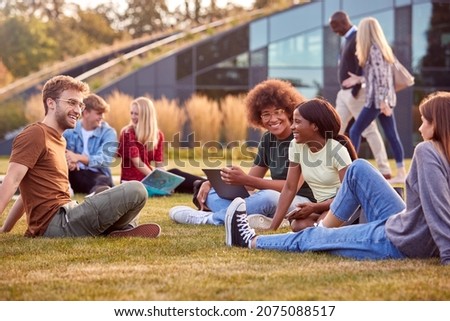 Group Of University Or College Students Sit On Grass Outdoors On Campus Talking And Working