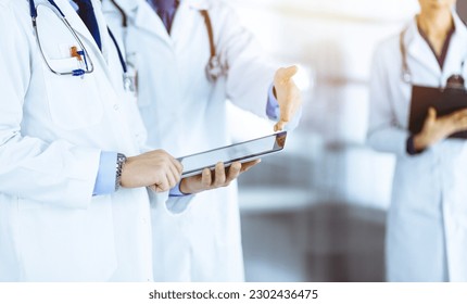Group of unknown doctors use a computer tablet to check up some medical names records, while standing in a sunny hospital office. Physicians ready to examine and help patients - Shutterstock ID 2302436475