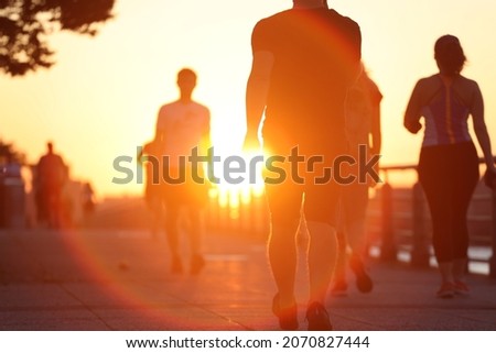 Group of unrecognized people exercise active walking on riverside boardwalk at sunset. Summer outdoor commuting fitness concept. 